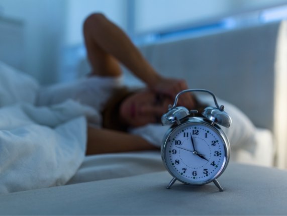 Person lying awake in bed next to nightstand with analog clock showing the time as 4 A M