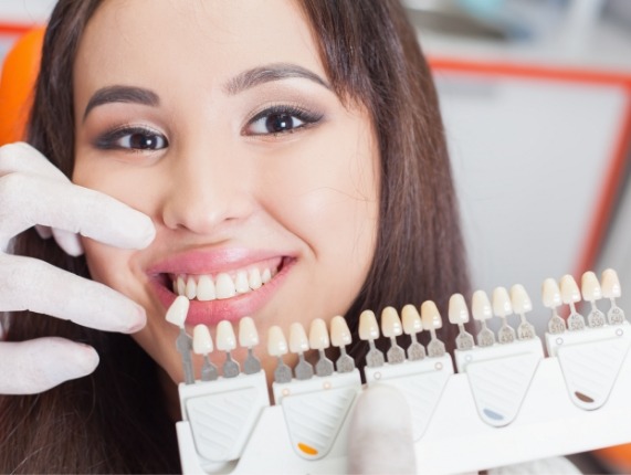 Young woman smiling while dentist holds shade guide in front of her smile